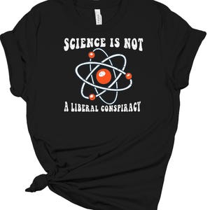 May include: Black t-shirt with the white text "SCIENCE IS NOT A LIBERAL CONSPIRACY." An atom graphic with white orbital paths and a red nucleus is centered above the text. The shirt is made of a soft material.
