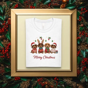 May include: White t-shirt featuring four dachshunds in festive Christmas attire, including Santa hats and reindeer antlers. The phrase "Merry Christmas" is written in red script below the dogs. The shirt is displayed in a gold frame with a holiday wreath.
