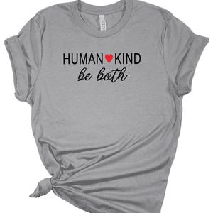 May include: A light gray t-shirt with the words "HUMAN KIND" in black block letters, a red heart symbol, and the words "be both" in a cursive font. The shirt is knotted at the bottom.