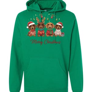 May include: Green hooded sweatshirt featuring a festive design. The graphic includes four dachshunds dressed in Christmas attire, with Santa hats, reindeer antlers, and sweaters. The words "Merry Christmas" are written in red script below the dogs.