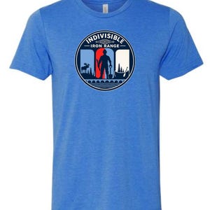 May include: A heather blue short-sleeve t-shirt featuring a circular graphic on the chest. The graphic displays the text INDIVISIBLE IRON RANGE, a silhouette of a person holding a shovel, a moose, pine trees, and a canoe against a red and white background.
