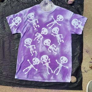 May include: Purple t-shirt with a white bleach design of skeletons dancing.