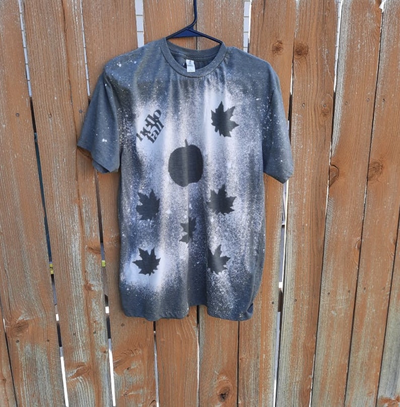May include: A dark gray t-shirt with a bleach-splattered design featuring a pumpkin and maple leaves. The text "Hello Fall" is written on the shirt.