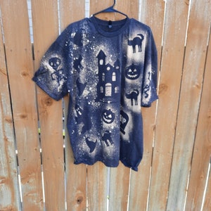 May include: A black t-shirt with a bleach-splattered design featuring various Halloween-themed silhouettes, including a haunted house, pumpkins, ghosts, and cats.