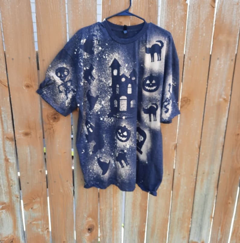 May include: A black t-shirt with a bleach-splattered design featuring various Halloween-themed silhouettes, including a haunted house, pumpkins, ghosts, and cats.