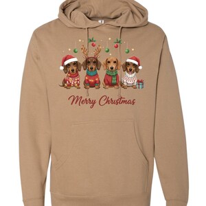 May include: A tan hoodie featuring a festive design with four dachshunds dressed in Christmas attire. Each dog wears a different outfit, including Santa hats and sweaters. The words "Merry Christmas" are printed in red.
