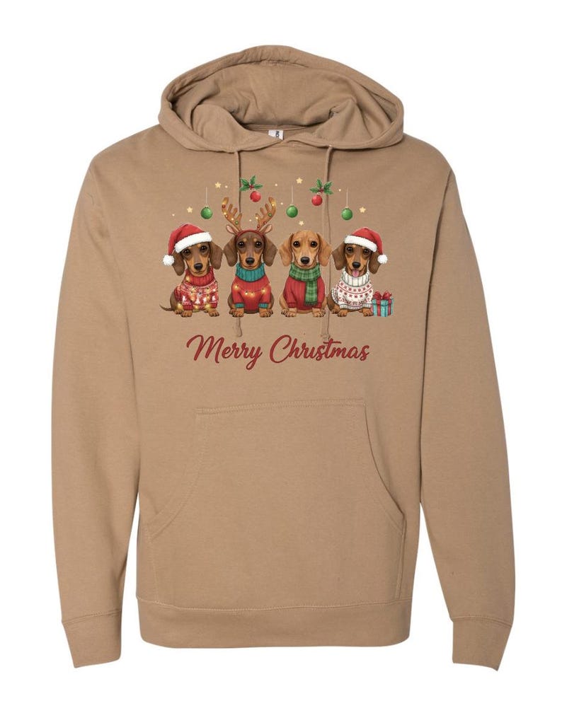 May include: A tan hoodie featuring a festive design with four dachshunds dressed in Christmas attire. Each dog wears a different outfit, including Santa hats and sweaters. The words "Merry Christmas" are printed in red.