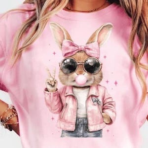 Könnte beinhalten: Pinkes T-Shirt mit einem Motiv eines Cartoon-Kaninchens, das eine Sonnenbrille, eine rosa Jacke und eine rosa Schleife trägt. Das Kaninchen zeigt ein Peace-Zeichen und hat eine Blase mit rosa Kaugummi im Mund.