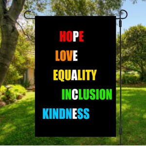 Könnte beinhalten: Eine schwarze Gartenflagge mit den Wörtern "HOPE", "LOVE", "EQUALITY", "INCLUSION" und "KINDNESS" in Rot, Orange, Gelb, Grün und Blau. Die Flagge hängt in einem Garten mit grünem Gras und Bäumen.