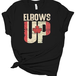 Puede incluir: Camiseta negra con el texto "Elbows Up" en letras blancas. El "Up" está estilizado como una bandera canadiense con una hoja de arce roja.