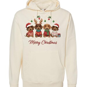 May include: Cream-colored hooded sweatshirt featuring a graphic of four dachshunds dressed in Christmas attire. The dogs wear Santa hats, reindeer antlers, and sweaters. The words "Merry Christmas" are printed in red below the dogs.