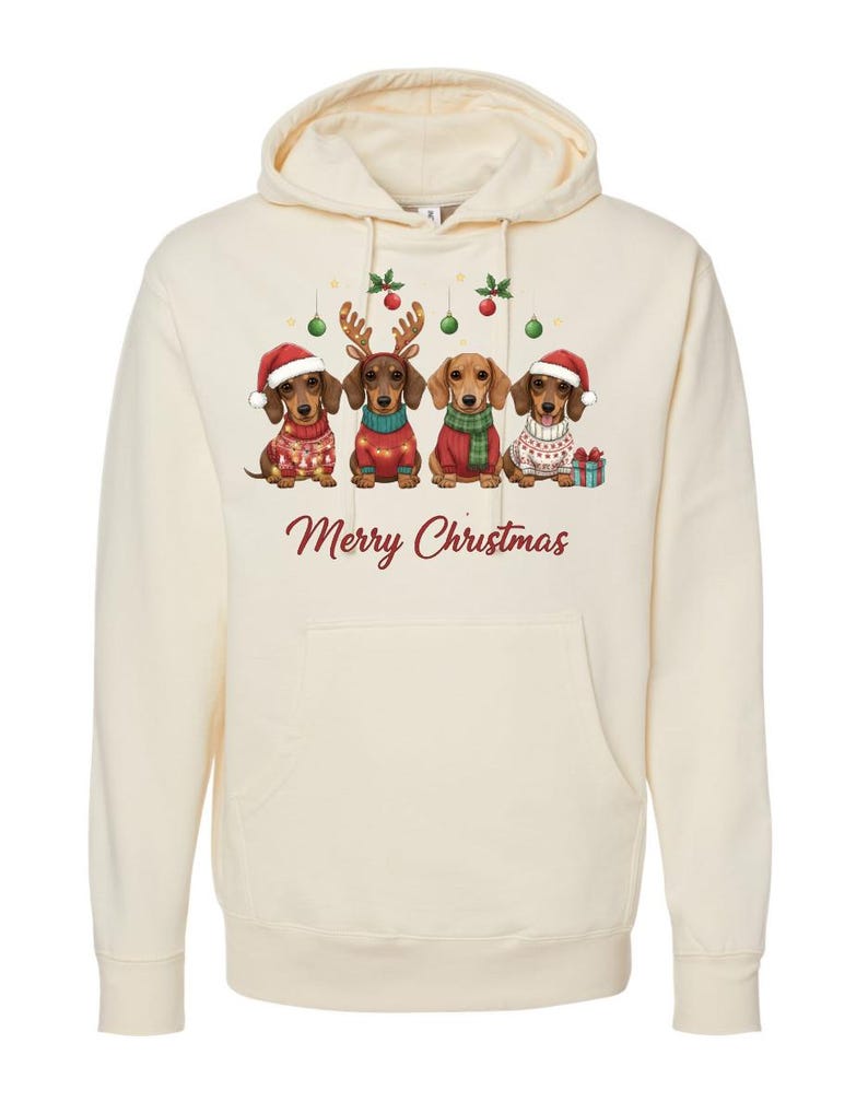 May include: Cream-colored hooded sweatshirt featuring a graphic of four dachshunds dressed in Christmas attire. The dogs wear Santa hats, reindeer antlers, and sweaters. The words "Merry Christmas" are printed in red below the dogs.