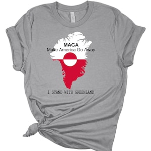 Puede incluir: Camiseta gris con un contorno blanco de Groenlandia con la bandera de Groenlandia superpuesta. El texto "Maga Make America Go Away" está por encima del mapa y "I Stand With Greenland" está por debajo del mapa.