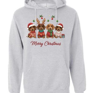 May include: A light gray hoodie featuring a graphic of four dachshunds dressed in Christmas attire. The dogs wear Santa hats, reindeer antlers, and scarves. The text "Merry Christmas" is printed in a festive font.