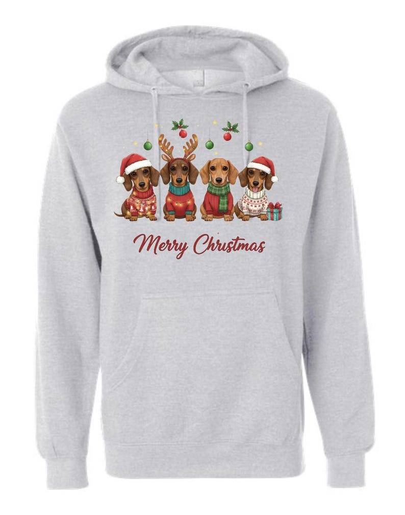 May include: A light gray hoodie featuring a graphic of four dachshunds dressed in Christmas attire. The dogs wear Santa hats, reindeer antlers, and scarves. The text "Merry Christmas" is printed in a festive font.