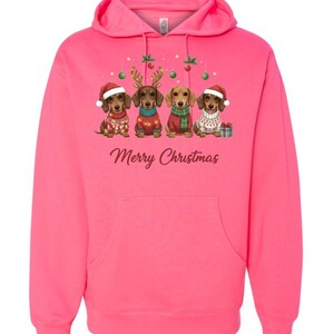 May include: A bright pink hoodie featuring a festive design. The graphic shows four dachshunds dressed in holiday attire, including Santa hats and sweaters, with the text "Merry Christmas" below. The hoodie has a front pocket and a hood.