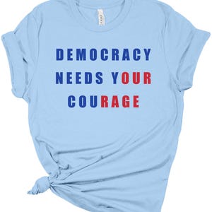 May include: Light blue t-shirt with the message "DEMOCRACY NEEDS YOUR COURAGE" printed in blue and red block letters. The shirt is made of a soft material and has a classic crew neck design.