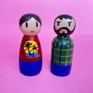May include: Two wooden peg dolls, one with brown hair and a red shirt with a palm tree design, the other with a brown beard and a green plaid shirt. Both dolls have blue pants.