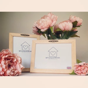 Può includere: Due cornici per foto in legno con sfondo bianco e una casa con un motivo a forma di cuore. Il testo "MYuniqueDECOR" è stampato su ogni cornice. Le cornici sono decorate con accenti metallici decorativi. Sullo sfondo ci sono fiori rosa.