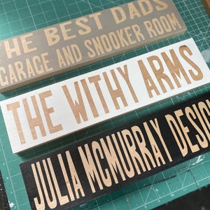 Könnte beinhalten: Drei Holzschilder mit goldener Beschriftung. Das oberste Schild ist grau und trägt die Aufschrift "The Best Dads Garage and Snooker Room". Das mittlere Schild ist weiß und trägt die Aufschrift "The Withy Arms". Das unterste Schild ist schwarz und trägt die Aufschrift "Julia McMurray Design".