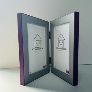 May include: A double photo frame with a purple wood border and a silver frame. The frame is hinged and has a house with a heart design on each side. The text "MY UNIQUE DECOR" is printed on each side of the frame.