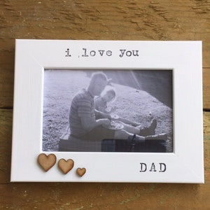 May include: White photo frame with the words "i love you" and "DAD" printed on it. The frame has a black and white photo of a father and child sitting on a grassy hill. There are three small wooden hearts on the bottom of the frame.