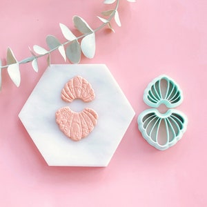Puede incluir: Dos formas de concha de fondant rosa sobre una baldosa de mármol blanco, junto a un cortador de galletas de concha de plástico verde.