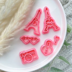 Könnte beinhalten: Rosa Plastik-Ausstecher in Form des Eiffelturms, einer Sonnenbrille, eines Vorhängeschlosses, eines Schlüssels und einer Kamera. Die Eiffelturm-Ausstecher haben ein Herz mit dem Wort "love" darin.