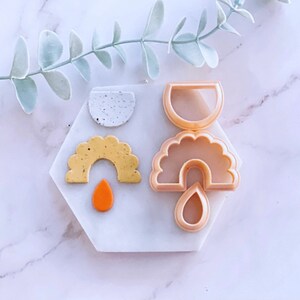 May include: A set of four cookie cutters in the shape of a rainbow, a teardrop, a half-circle, and a speckled circle. The cutters are made of a light beige plastic and are displayed on a white hexagonal tile.