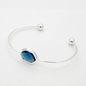 May include: A silver-toned cuff bracelet with a faceted, deep blue gemstone at its center. The open-ended design features small, round accents at each end. The gemstone is an octagonal shape.