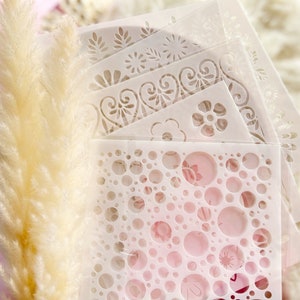 May include: A set of six white plastic stencils with various designs, including circles, flowers, and swirls. The stencils are stacked on top of each other and are sitting on a white fluffy surface.