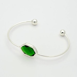 May include: A silver-toned cuff bracelet featuring a vibrant green, emerald-cut gemstone at its center. The open design has a small silver ball at each end. The gemstone is set in a silver bezel, creating a modern and elegant look.