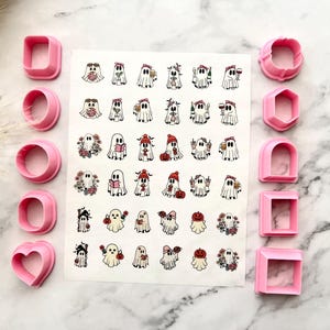 Può includere: Un foglio di adesivi con varie illustrazioni di fantasmi disposte in file, affiancato da dieci tagliabiscotti in plastica rosa in varie forme geometriche, tra cui cerchi, quadrati, un cuore, un esagono e un arco su una superficie di marmo.