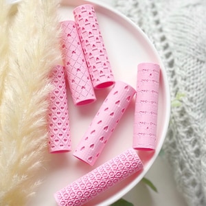 May include: Six pink hair rollers with different embossed patterns, including hearts, diamonds, and XOXO. The rollers are on a white surface.