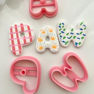 Puede incluir: Tres cortadores de galletas de plástico rosa con forma de las letras P, A y W, junto a tres galletas decoradas con glaseado blanco. La P tiene una cuadrícula rosa, la A huellas de patas naranjas y la W motas azules y verdes.