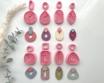 May include: A set of 12 pink plastic earring molds in various shapes, including circles, ovals, teardrops, and squares. The molds are arranged in three rows of four. Each mold has multiple concentric rings, creating a layered effect.