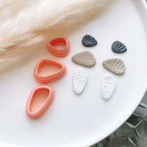 Op de afbeelding: Drie roze plastic bladeren koekjesvormen en zes polymer clay bladeren in verschillende tinten bruin, wit en zwart.