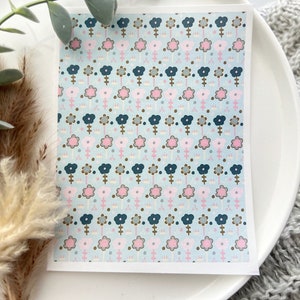 May include: A rectangular card with a repeating floral pattern. The flowers are in rows and columns, with alternating colors of pink, navy, and gray on a light blue background. The card is on a white plate.