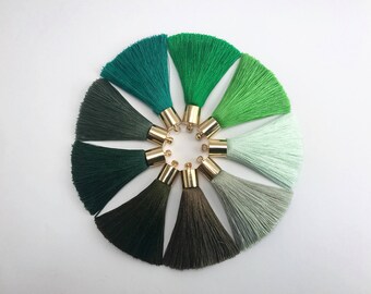 Puede incluir: Un conjunto de diez borlas de diferentes tonos de verde. Las borlas están dispuestas en círculo, con los anillos metálicos dorados en la parte superior de cada borla mirando hacia adentro.