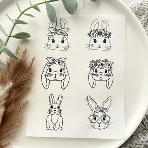 May include: A white paper with six black and white bunny illustrations. The bunnies have different accessories, including bows, flower crowns, and glasses. The image is on a white plate with greenery and a gray knitted blanket in the background.