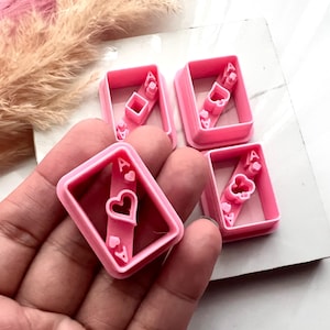 Puede incluir: Cuatro cortadores de galletas de plástico rosa con forma de As de corazones. Los cortadores tienen un marco rectangular con el símbolo del corazón y la letra "A" visible. Los cortadores están sobre una superficie blanca.
