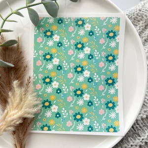 May include: A floral patterned card featuring a repeating design of teal, white, yellow, and pink flowers on a light green background. The card is displayed on a white plate with decorative dried plants.