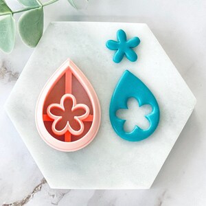 Puede incluir: Dos cortadores de galletas de plástico rosa y azul con forma de lágrima con un diseño de flor en el interior. También se incluye un cortador de galletas de flor rosa más pequeño.