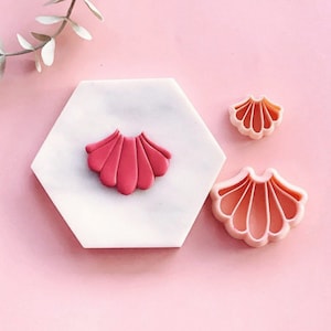 Puede incluir: Tres cortadores de galletas con forma de concha de plástico rosa y blanco sobre una superficie hexagonal blanca. El cortador más grande es rosa y los dos más pequeños son blancos.