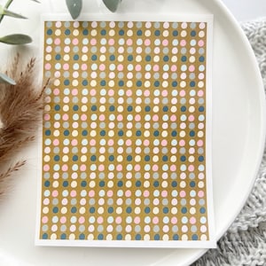 May include: A rectangular card with a pattern of small, evenly spaced dots in pastel colors of pink, blue, and white on a gold background. The card is set on a white plate, with dried plants and a gray knitted blanket in the background.