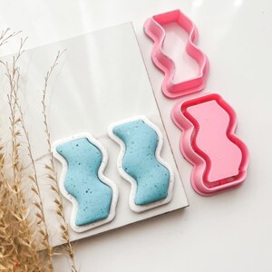 Puede incluir: Dos pendientes de arcilla polimérica de forma ondulada azul y blanca y dos moldes de plástico rosa para hacer los pendientes.