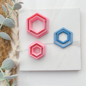 Könnte beinhalten: Drei sechseckige Ausstecher, zwei rosa und einer blau. Die rosa Ausstecher sind ineinander verschachtelt, wobei der größte Ausstecher oben liegt. Der blaue Ausstecher befindet sich auf der rechten Seite des Bildes.