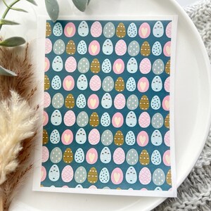 May include: A greeting card with a teal background and a pattern of Easter eggs in various colors and designs, including pink hearts, gold dots, and blue stripes. The card is on a white surface with decorative elements.