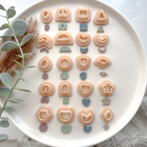 May include: A set of 20 small, peach-colored polymer clay shapes on a white plate. The shapes include a heart, star, moon, and various geometric shapes.