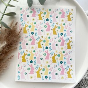 May include: A white sticker sheet with a repeating pattern of yellow and pink bunnies, blue and yellow flowers, and green leaves. The sticker sheet is on a white plate.